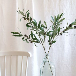 May include: A green olive branch with black olives, arranged in a clear glass vase. The vase is on a white surface.