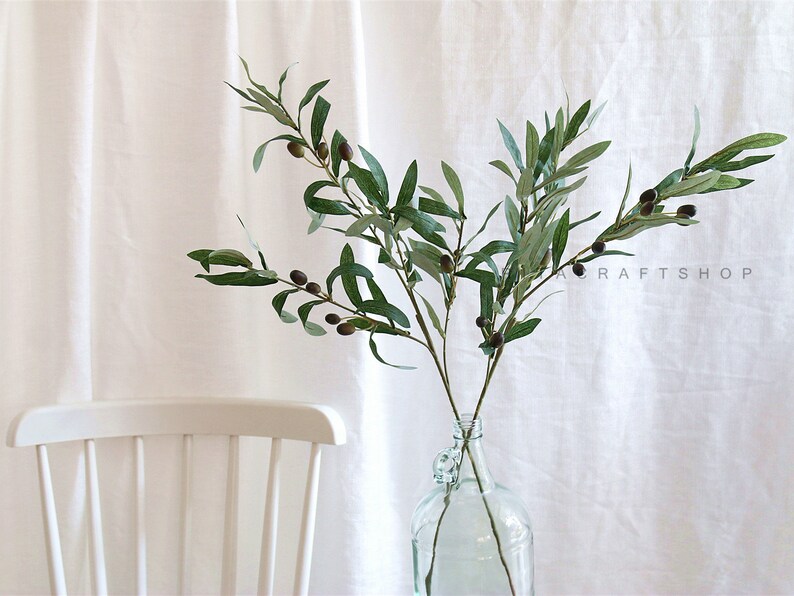 May include: A green olive branch with black olives, arranged in a clear glass vase. The vase is on a white surface.