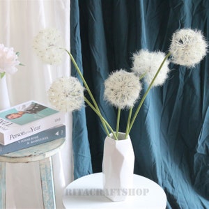 Peut inclure: Cinq tiges de pissenlit blanc dans un vase géométrique blanc sur une table blanche. Le vase est sur une table blanche avec un fond bleu sarcelle.