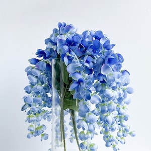 Pu&ograve; includere: Un vaso di vetro pieno di un mazzo di fiori artificiali blu. I fiori sono disposti a cascata, con gli steli che si estendono lungo i lati del vaso.
