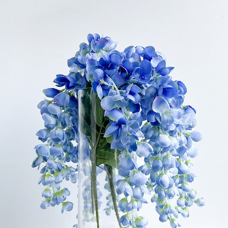Pu&ograve; includere: Un vaso di vetro pieno di un mazzo di fiori artificiali blu. I fiori sono disposti a cascata, con gli steli che si estendono lungo i lati del vaso.