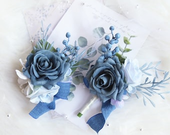Puede incluir: Dos boutonnières florales azules y blancas con detalles de cinta azul. Las boutonnières presentan una rosa azul, flores blancas y bayas azules.