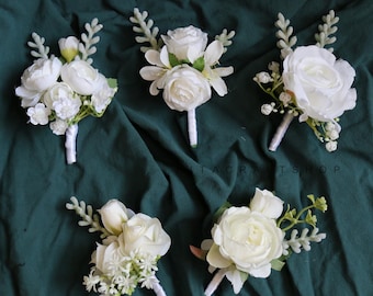 Pode incluir: Cinco botões florais brancos em tecido verde escuro. Cada botão apresenta diferentes flores brancas e vegetação, com caules envoltos em fita branca. Ideais para casamentos ou eventos especiais.