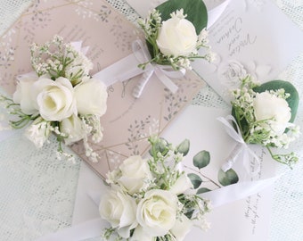 Pode incluir: Quatro boutonnières de rosas brancas com folhagem e fita branca. As boutonnières estão dispostas sobre uma superfície branca com convites de casamento.