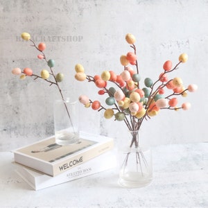 以下が含まれることがあります： パステルカラーの斑点のある卵が付いた人工の枝のブーケ。枝は、積み重ねられた2冊の本のあるテーブルの上の透明なガラスの花瓶に入っています。上の本には「WELCOME」という文字が表紙に印刷されています。