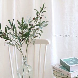 May include: A green olive branch with dark green leaves and small black olives in a clear glass vase. The vase is sitting on a white wooden chair. The background is a white wall with the text "RITACRAFTSHOP" printed on it.