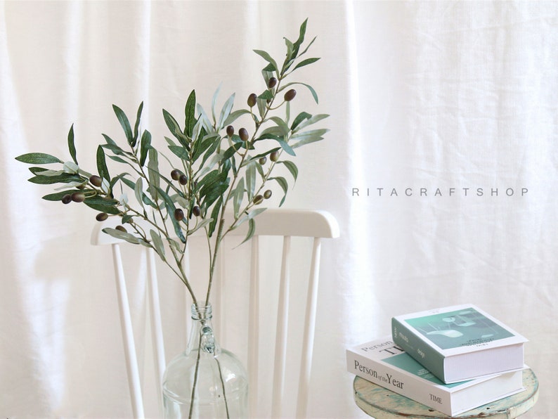 May include: A green olive branch with dark green leaves and small black olives in a clear glass vase. The vase is sitting on a white wooden chair. The background is a white wall with the text "RITACRAFTSHOP" printed on it.