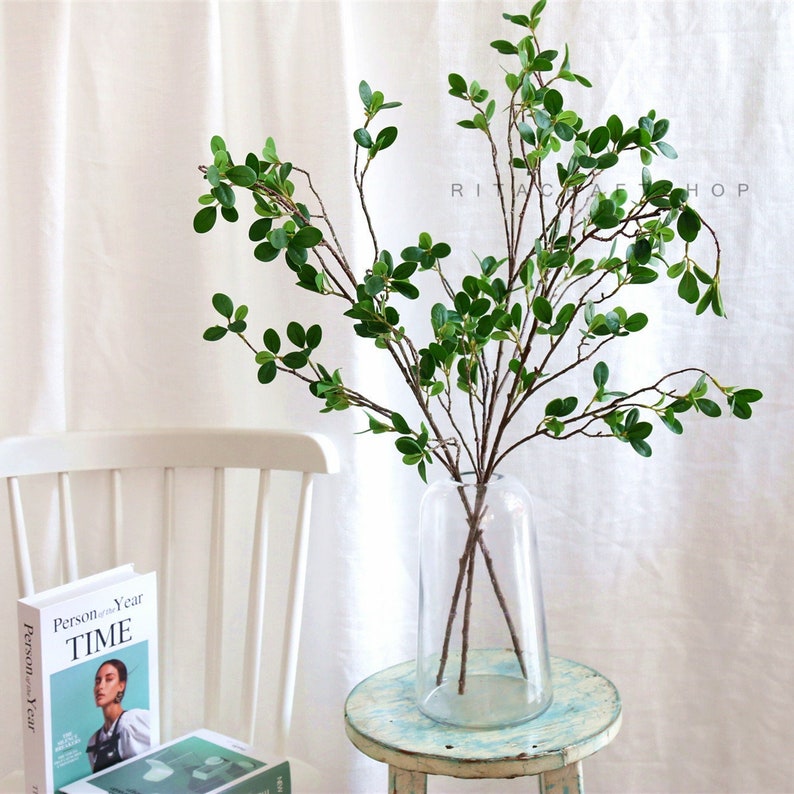 May include: A clear glass vase with a wide mouth and a narrow base holds a bouquet of artificial green leafy branches. The vase is sitting on a light blue wooden table.
