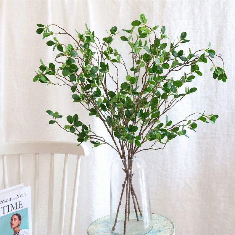 May include: A bouquet of artificial green leafy branches in a clear glass vase. The vase is sitting on a white surface.