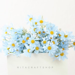 May include: A bouquet of light blue artificial daisies with yellow centers. The flowers are arranged in a white square container. The text "RITACRAFTSHOP" is printed at the bottom of the container.