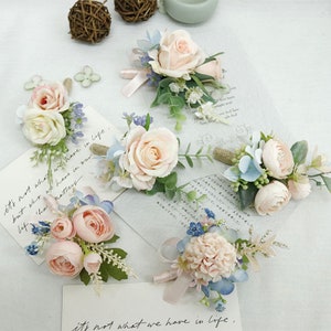 May include: Five artificial flower boutonnieres with pink and blue flowers. The boutonnieres are arranged on a white surface with text that reads "it's not what we have in life, but who we have in our life matters".