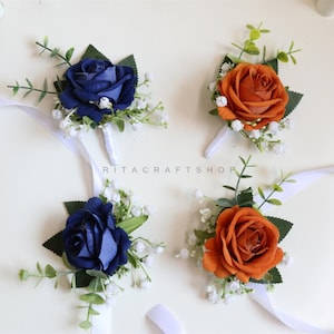 Könnte beinhalten: Vier florale Handgelenks-Corsagen. Zwei sind marineblaue Rosen und zwei sind orangefarbene Rosen. Jede Corsage hat ein weißes Band, grüne Blätter und kleine weiße Blüten. Der Text "RITA CRAFT SHOP" ist sichtbar.