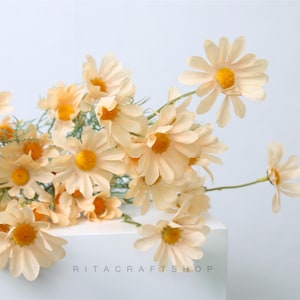 Può includere: Un mazzo di margherite artificiali color crema con centri gialli. I fiori sono disposti in uno stile sciolto e naturale e hanno un aspetto delicato e vintage.