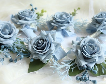 Puede incluir: Seis boutonnières de rosa artificial de color azul claro con hojas verdes y bayas azules. Las boutonnières están dispuestas sobre un fondo de tela blanca.