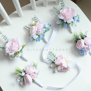 May include: A collection of handmade floral boutonnieres and wrist corsages. Each piece features soft pink roses and smaller blue and white accent flowers, with green foliage. Some have white ribbon streamers. The shop name "RITACRAFTSHOP" is visible.