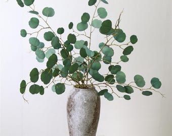 May include: A vase filled with artificial eucalyptus branches with green leaves. The vase is a light gray color with a textured surface. The vase is sitting on a white surface. The text "RITA CRAFTS SHOP" is visible at the bottom of the image.