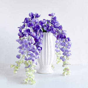Pu&ograve; includere: Un vaso di ceramica bianco con una superficie testurizzata contiene un bouquet di fiori di glicine viola. I fiori cadono a cascata sui lati del vaso.