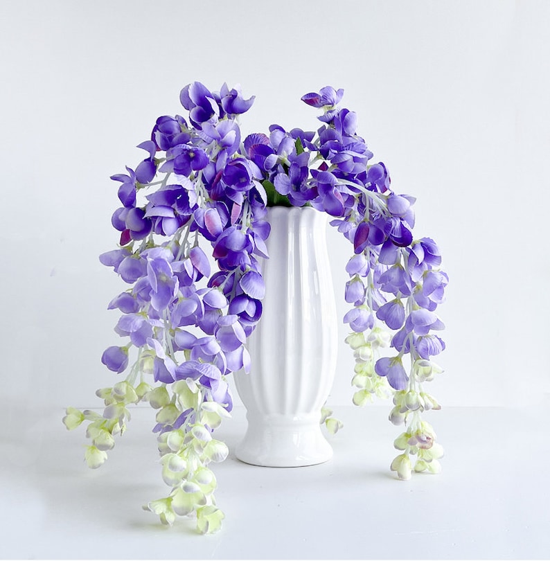 Pu&ograve; includere: Un vaso di ceramica bianco con una superficie testurizzata contiene un bouquet di fiori di glicine viola. I fiori cadono a cascata sui lati del vaso.