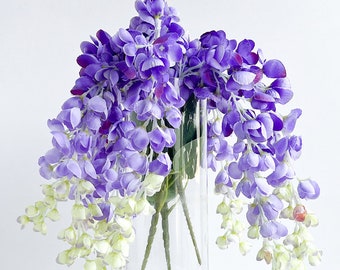May include: A bouquet of purple and white wisteria flowers in a clear glass vase. The flowers are arranged in a cascading style, with the purple blooms hanging down from the vase.