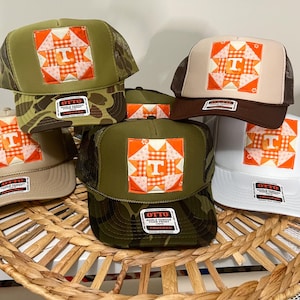 May include: A collection of trucker hats with a square patch featuring a quilt-like design in orange, white, and peach. The hats have various color schemes, including khaki, brown, green, and white, with mesh backs and the letter "T".