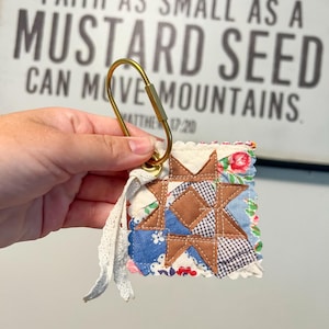 May include: A square quilted keychain featuring a brown star pattern on a patchwork background of blue, white, and floral fabrics. The keychain is attached to a gold-toned metal carabiner and includes a decorative white lace ribbon accent.