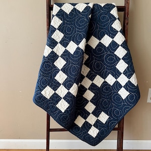 May include: A navy blue and white quilt with a diamond pattern is draped over a wooden ladder. The quilt features a repeating pattern of white diamonds on a navy blue background. The quilt is quilted with a swirling pattern.