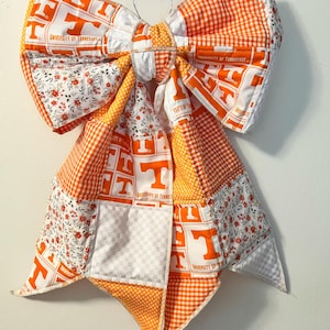 May include: A large, handmade fabric bow featuring orange and white patchwork. The bow incorporates various patterns, including gingham, floral, and fabric printed with "UNIVERSITY OF TENNESSEE" and the letter "T". It is hanging on a metal hanger.