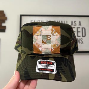 May include: A camouflage trucker hat with a patchwork square design in the center. The patch features floral and geometric patterns in shades of pink, yellow, and green. The hat has a curved brim and a mesh back. The label reads "OTTO TRUCKER".