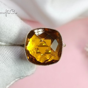 May include: A silver ring featuring a large, square-cut, amber-coloured gemstone. The gemstone is multi-faceted, catching the light. The ring is displayed against a pink backdrop with white fabric and foliage. The brand name "Gemscrafts Co" is visible.