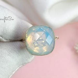 May include: A square-cut opal ring with a silver band. The opal has a light blue and white iridescent appearance. The ring is set against a pink background.