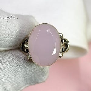 May include: A silver ring with a large, oval, pale pink gemstone. The gemstone is faceted and set in a decorative silver band. The ring is displayed on a white surface with a pink background. The text "Gemserafts Co" is visible.