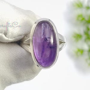 May include: A silver ring featuring a large, oval-shaped, smooth purple amethyst gemstone set in a polished metal bezel. The band has a split design on the sides, and the text GemcraftsCo is visible in the upper left corner of the image.