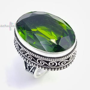 May include: A silver ring with a large, oval-shaped, vivid green gemstone. The ring has an intricate, patterned band. The gemstone is the main feature, displaying a rich, green colour. The ring is set against a white background. The ring is a piece of jewellery.