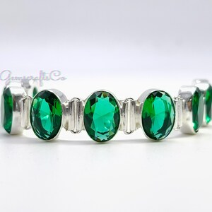 May include: A silver-toned link bracelet featuring a series of oval-shaped, faceted emerald green stones. Each stone is securely set within a polished metal frame, connected by small, sturdy silver-colored metal links to form a continuous chain.