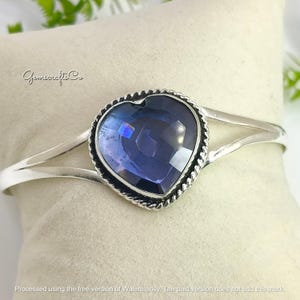May include: A silver-toned cuff bracelet featuring a large, heart-shaped, faceted, deep blue gemstone. The gemstone is set within a twisted rope-like border. The bracelet has a split design on each side of the heart.