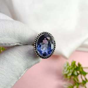 May include: A silver-toned ring with an oval, faceted, blue gemstone. The gemstone is surrounded by a twisted rope-like border. The ring is set against a soft pink background with white fabric and green foliage.
