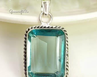 May include: A rectangular pendant with a large, light blue gemstone set in a silver-colored metal frame. The frame has a twisted rope design around the gemstone. The pendant has a loop at the top for a chain. The text "Gemserafts Co" is visible.