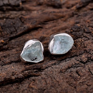 Pode incluir: Um par de brincos de pedra aguamarina com moldura de prata. Os brincos têm uma forma natural e irregular e uma cor azul pálido. O fundo é uma superfície texturizada de cor castanho escuro.