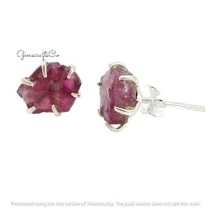 May include: A pair of stud earrings with rough-cut, deep pink gemstones set in silver-coloured metal. The gemstones have a natural, irregular shape. The earrings are displayed against a white background.