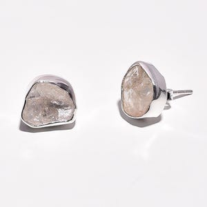 Puede incluir: Un par de pendientes de botón con piedras preciosas en bruto de color claro engastadas en metal plateado. Los pendientes tienen una forma natural y orgánica, con texturas visibles y variaciones.