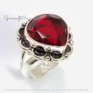 May include: A silver ring with a large, faceted, heart-shaped, deep red gemstone. The gemstone is set in an ornate silver setting with detailed design. The ring has a wide band.
