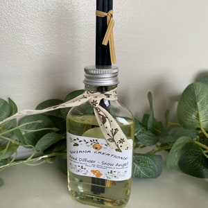 May include: A clear glass bottle with a silver lid and a white label that reads "Dhyana Creations Reed Diffuser - Snow Angel". The bottle is filled with a yellow liquid and has black reeds sticking out of the top. The bottle is tied with a white and yellow ribbon.
