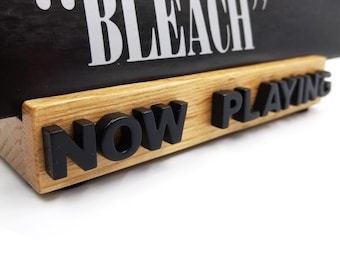 Peut inclure: Un support de présentation en bois avec le texte noir en relief NOW PLAYING sur le devant. Un panneau rectangulaire noir avec le texte blanc BLEACH est inséré dans la fente supérieure de la base en bois clair au grain naturel visible.