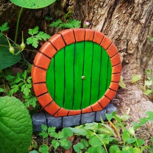 May include: A whimsical, round wooden door painted green with vertical black lines, framed by a brick-like orange border. The door is set against a tree trunk and surrounded by greenery, creating a fairy tale aesthetic.