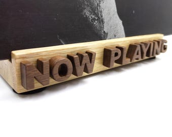 Peut inclure: Un support en bois clair comportant des lettres en relief marron foncé formant l'inscription NOW PLAYING. Ce support est conçu pour maintenir une pochette de disque vinyle noire, partiellement visible appuyée contre la base en bois.