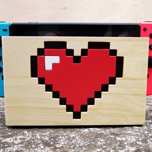 Peut inclure: Un support en bois avec un motif de cœur rouge pixelisé, tenant une console Nintendo Switch avec des Joy-Cons bleus et roses.
