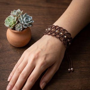 May include: A brown macrame bracelet with gold-colored beads is worn on a wrist. The bracelet has a woven design. A small terracotta pot with several succulents is to the left of the wrist. The background is a dark wooden surface.