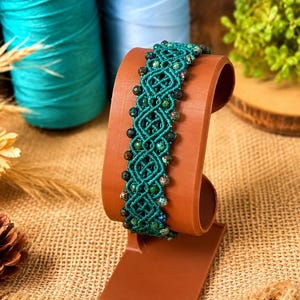 May include: A teal macrame bracelet with dark green and brown beads is displayed on a brown stand. The bracelet features a woven design with diamond-shaped patterns. The background includes spools of teal and light blue thread.