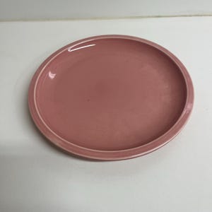 May include: A round, dusty rose-colored ceramic plate. The plate has a slightly raised rim and a smooth, glossy surface. The plate is designed for serving food.