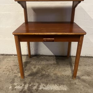 Puede incluir: Una mesa auxiliar de madera con un cajón y un estante. La mesa tiene un acabado marrón claro y está hecha de madera maciza.